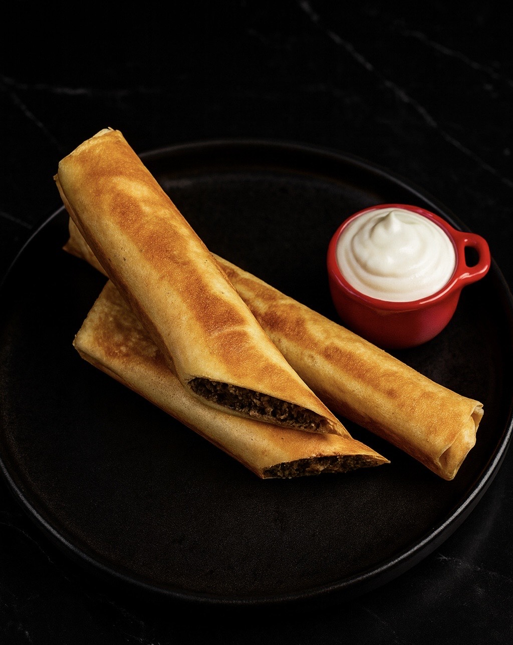 Savory meat-filled blinchiki rolls served on a black plate with a side of sour cream.